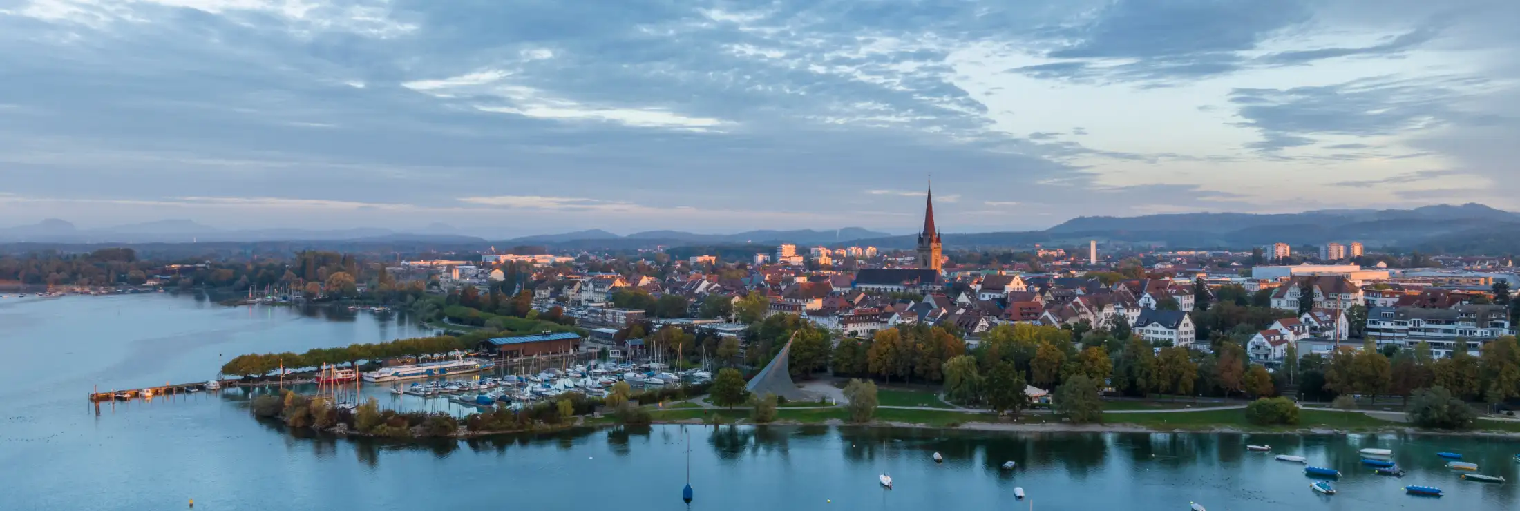 Luftaufnahme Radolfzell am Bodensee: links Hafenmole, Wäschbruckhafen und Konzertsegel, dahinter der Bahnhof, mittig Münster Unserer Lieben Frau und Kernstadt, rechts Immobilien Scheffelstraße und Kapuzinerweg, vorne der Bodensee mit einigen Booten.