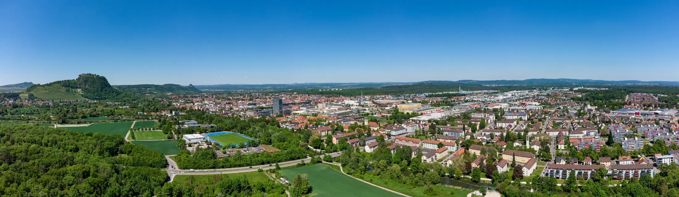 Luftaufnahme von Singen: links der Hohentwiel, vorne Münchriedstraße / Hombergerstr über die Radolfzeller Aach, dahinter Münchriedstadion, mittig die Kernstadt Singen, rechts Immobilien Lindenhain, weitere Immobilien südliche Kernstadt.