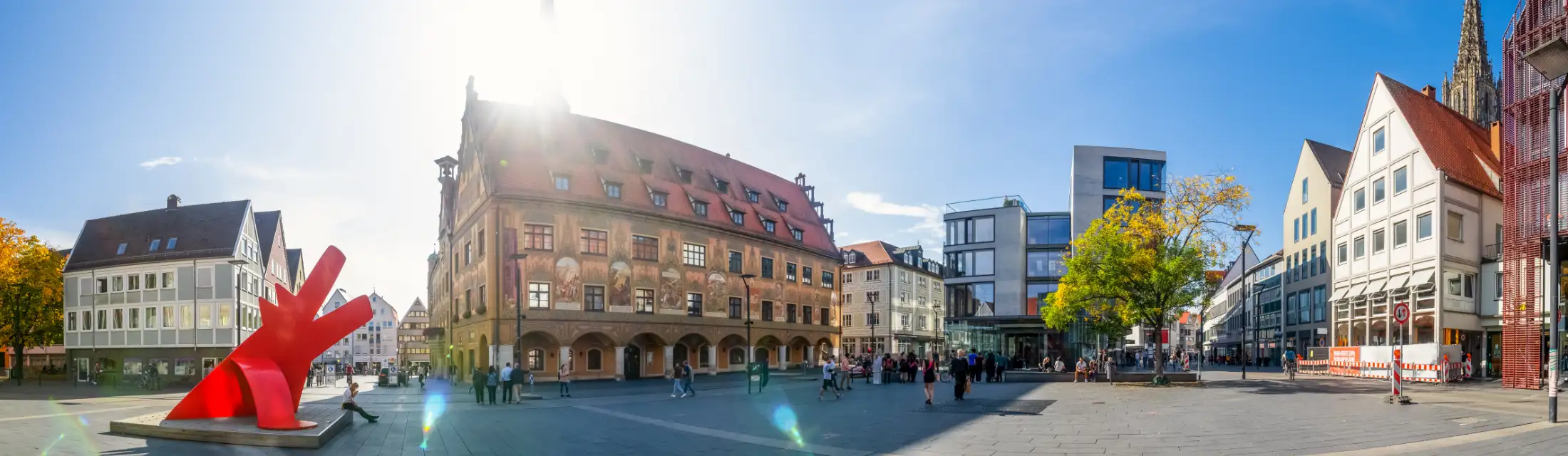 Panoramaaufnahme Markplatz Ulm: links Skulptur „Red Dog“ vor der Kunsthalle Weishaupt, Immobilien Neue Straße / südlicher Marktplatz, mittig Ulmer Rathaus, rechts Immobilien Hans und Sophie Scholl Platz / Neue Straße, dahinter Turm Ulmer Münster