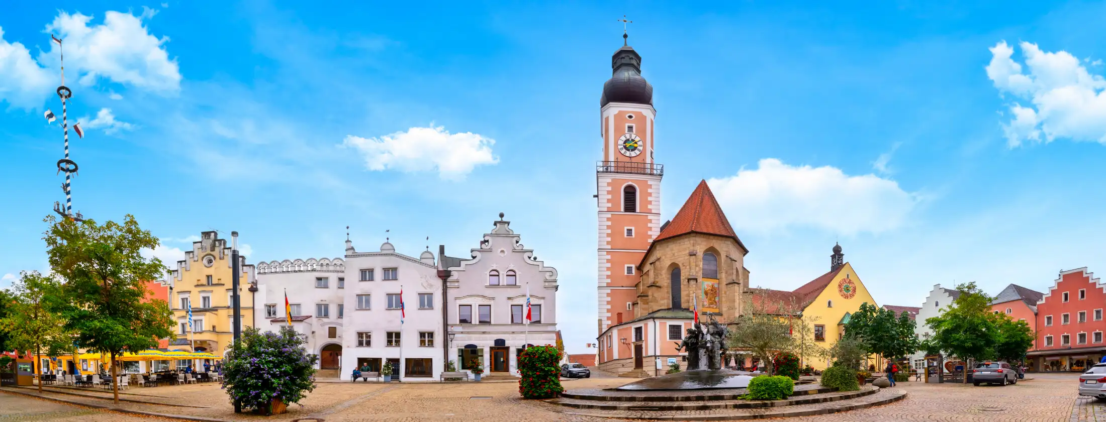 Der Marktplatz in Cham, im Vordergrund der Marktplatzbrunnen, dahinter die Stadtpfarrkirche St. Jakob, daneben weitere Immobilien des westlichen Marktplatzes, wie z. B. links Tourist-Info Cham und rechts das Rathaus am Marktplatz an einem Sommertag