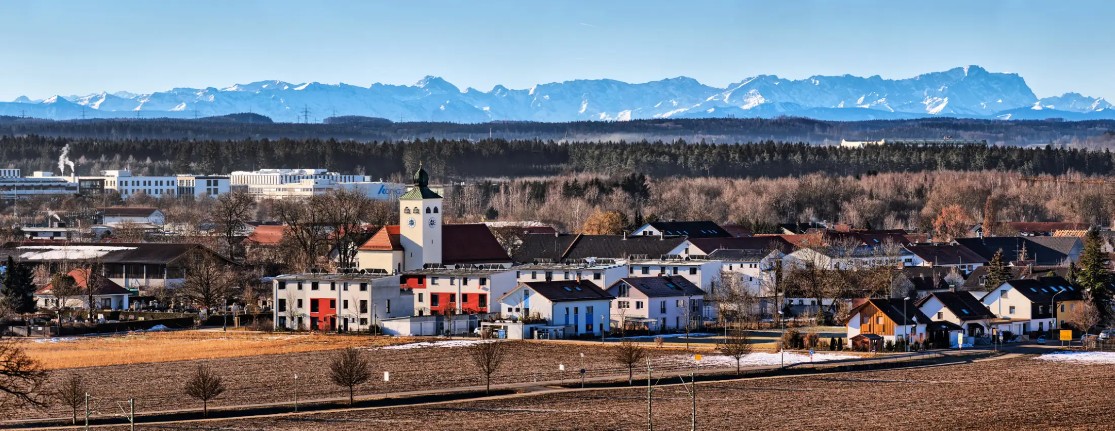 Panoramaaufnahme von Gilching: etwa mittig die Kirche St. Nikolaus in Gilching Argelsried, links dahinter die gewerblichen Immobilien Am Römerstein/Ferdinand Porsche Str., im Hintergrund die Alpen.