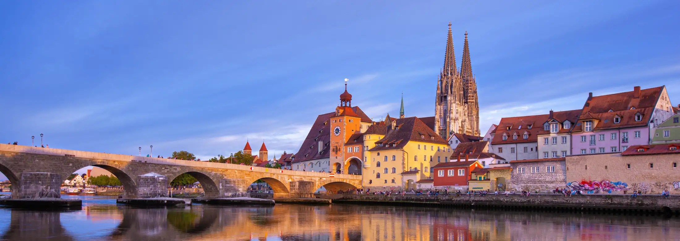 Panoramaaufnahme Dom St. Peter zu Regensburg: links der südliche Teil der Steinernen Brücke über die Donau, links die Türme Dompfarrkirche Niedermünster, mittig der Brückturm und Brückturm-Museum, rechts die Immobilien Am Wiedfang / Goldene-Bären Straße.