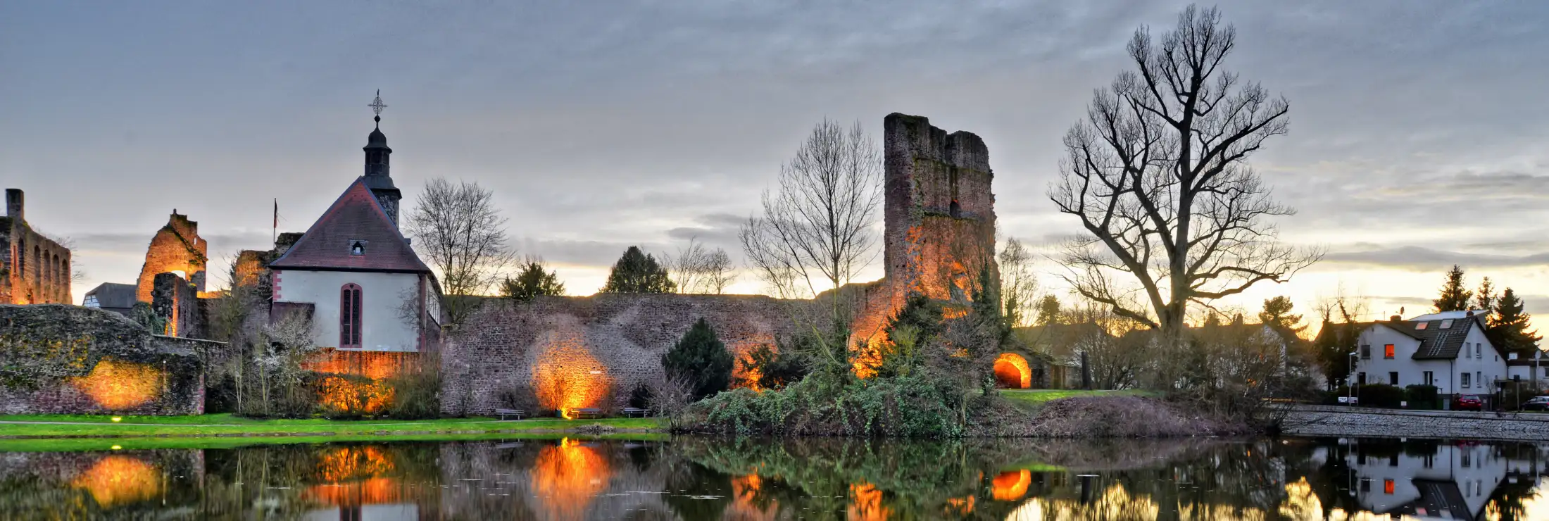Burg Hayn im Stadtteil Dreieichenhain in der Dämmerung. Im Vordergrund der Burgweiher, der die Beleuchtung reflektiert, links Immobilien Fahrgasse und Überreste des Palas, daneben die evangelische Burgkirche, rechts Immobilien Solmische Weiher Straße