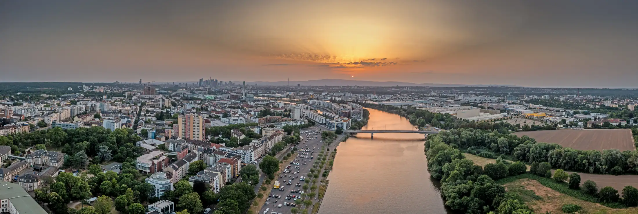 Luftbild von Offenbach am Main: links Büsing-Palais, Immobilien im Zentrum, Parkplatz Mainufer, dahinter Hafen und Kaiserlei, mittig Main und Carl-Ulrich-Brücke nach Frankfurt Fechenheim. Hinten Skyline Frankfurt Innenstadt und untergehende Sonne.