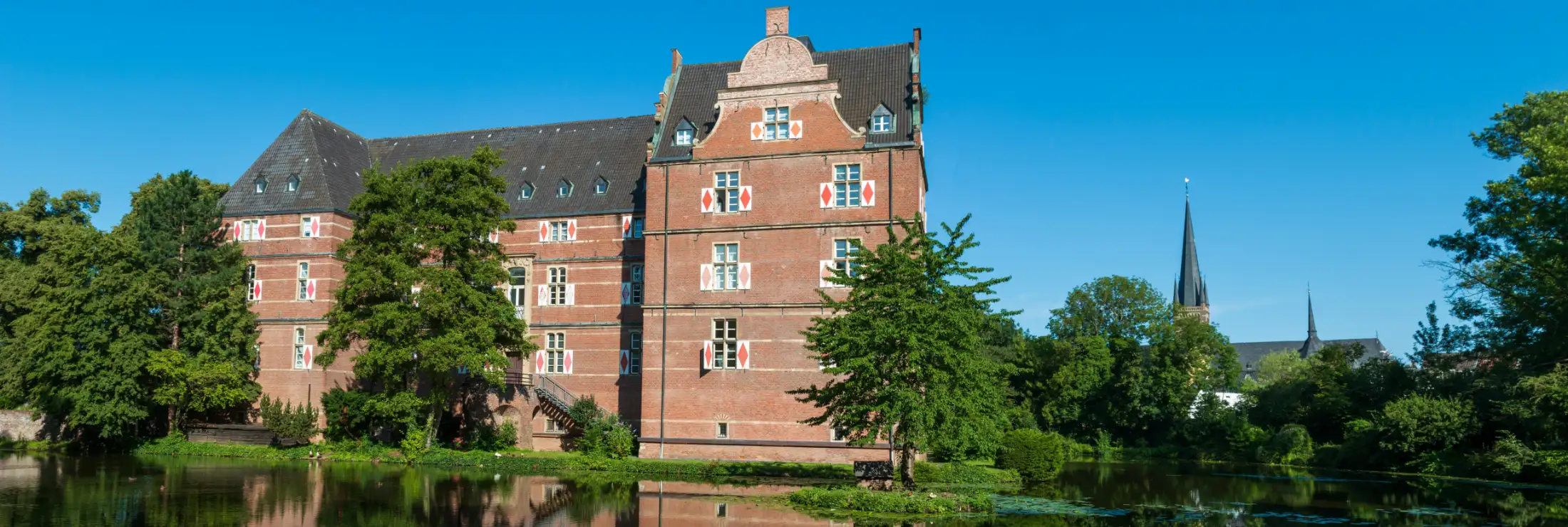 Schloss Bedburg: Aufgenommen aus dem Schlosspark, vorne der Wassergraben, links und mittig das Schloss Bedburg, rechts z. T. sichtbar die Kirche St. Lambertus.