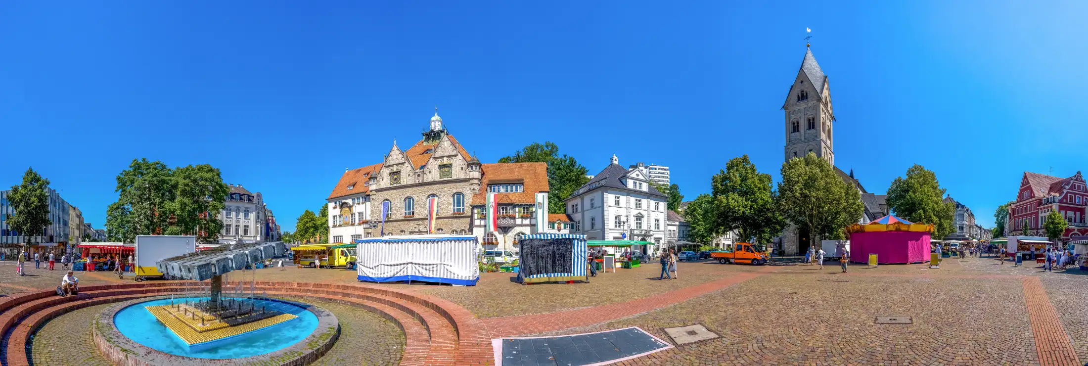 Panoramabild Markplatz am Brunnen Bergisch Gladbach: links Papierbrunnen, daneben Historisches Rathaus Stadtmitte und Heilige Figur, rechts Immobilie nördlicher Markplatz und Kirche St. Laurentius, ganz rechts Immobilien östlicher Marktplatz / Hauptstraße