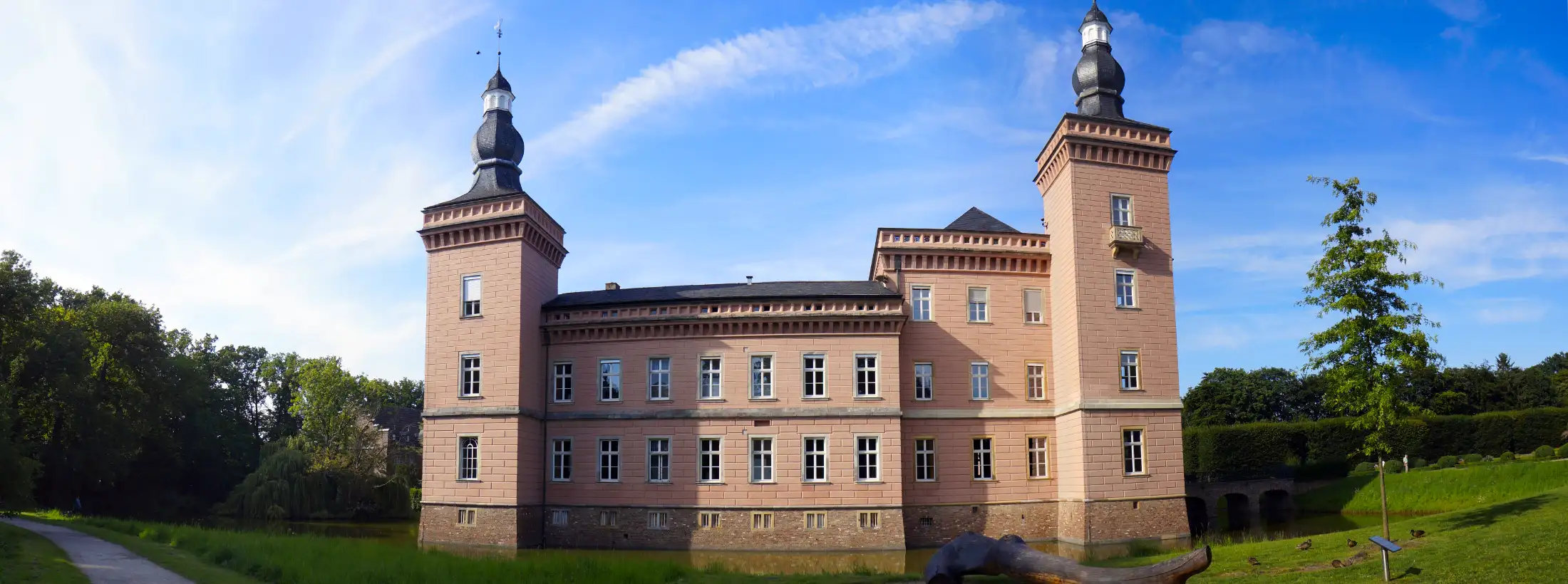 Schloss Gracht, ein Wasserschloss: mittig das Herrenhaus, umgeben vom Wassergraben, links und rechts der Schlosspark Gracht an einem sonnigen Tag.