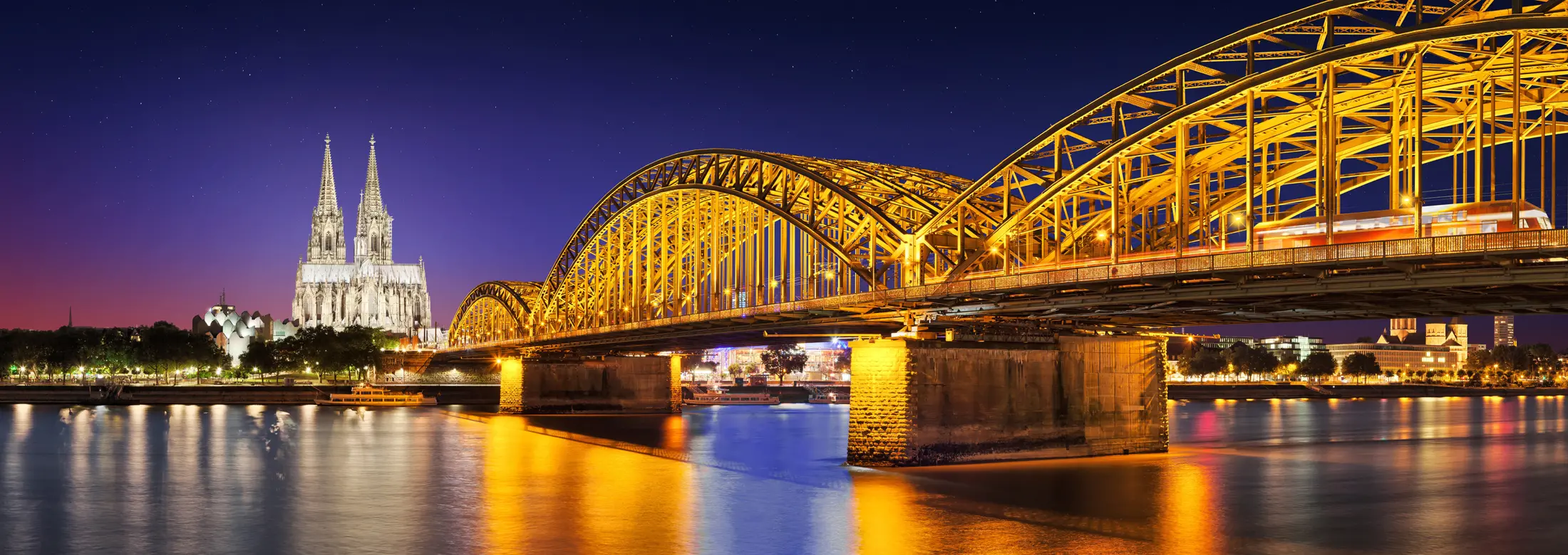 Köln bei Nacht, mit der Hohenzollernbrücke im Vordergrund und dem beleuchteten Köner Dom und vielen Immobilien am Rheinufer im Hintergrund