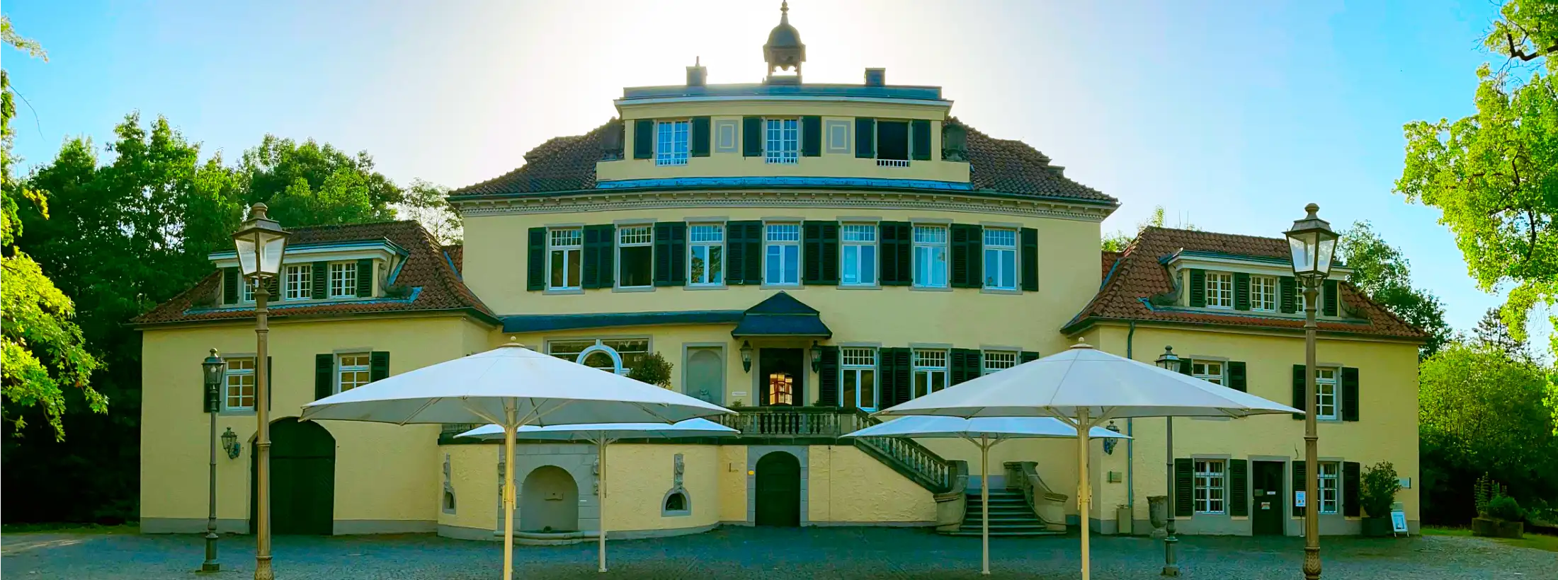 Bild von Schloss Eulenbroich, eine der bekanntesten Immobilien in und Wahrzeichen von Rösrath. Zu sehen ist das Herrenhaus, aufgenommen vom Torbogen aus, Blickrichtung Osten. Hinter dem Gebäude die aufgehende Sonne, an einem Sommermorgen