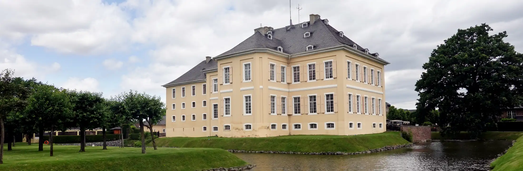 Das Barockschloss Miel in Swisttal von Süden: vorne der Wassergraben, links Bäume der Obstwiese Schloss Miel, mittig der Schlossbau, rechts Immobilien Schlossallee