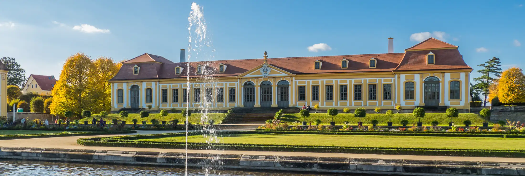 Aufnahme aus dem Barockgarten Großsedlitz von der Oberen Orangerie an einem Sommertag mit blauem Himmel, davor Wiesen, Blumen, Zierpflanzen und ein Springbrunnen bzw. Fontäne, links ist ein kleiner Teil des Friedrichschlösschen sichtbar.